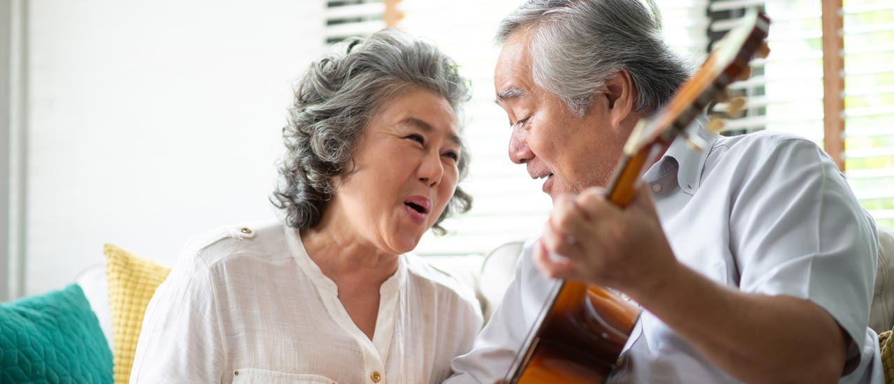 A gray haired man sits holding a guitar next to a gray haired woman who is singing along