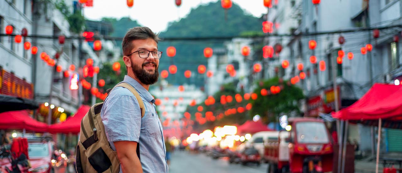 A bearded man with glasses and a backpack stands on a street with Chinese red lanterns hanging on wires above