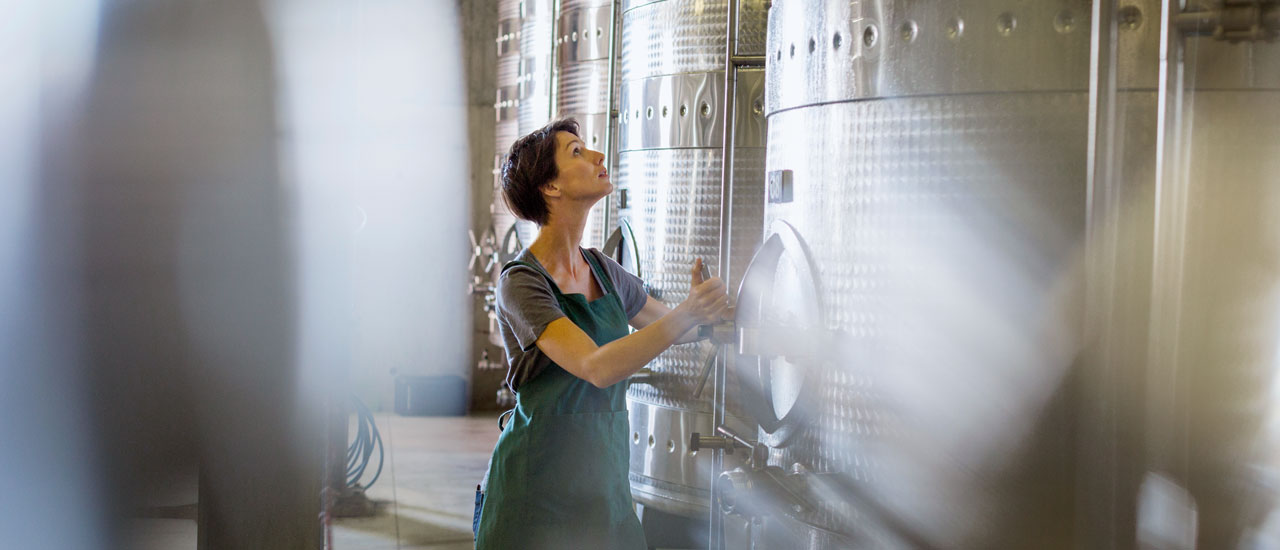 A woman looks up as she adjusts a stainless steel tank in a room full of large-scale brewing vats.