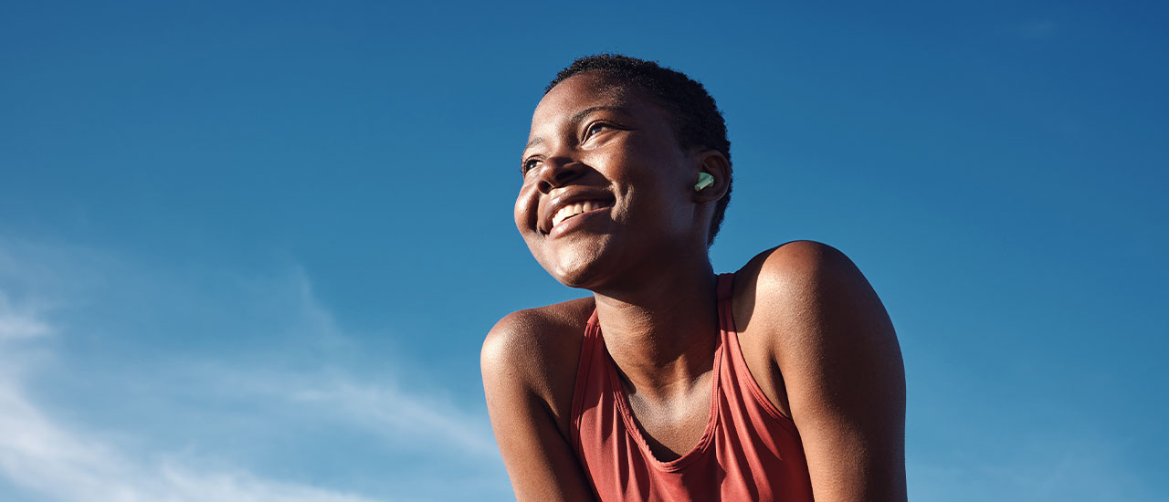 A woman in a sleeveless athletic top leans forward with arms braced on her knees, photographed from a low angle against a clear blue sky.