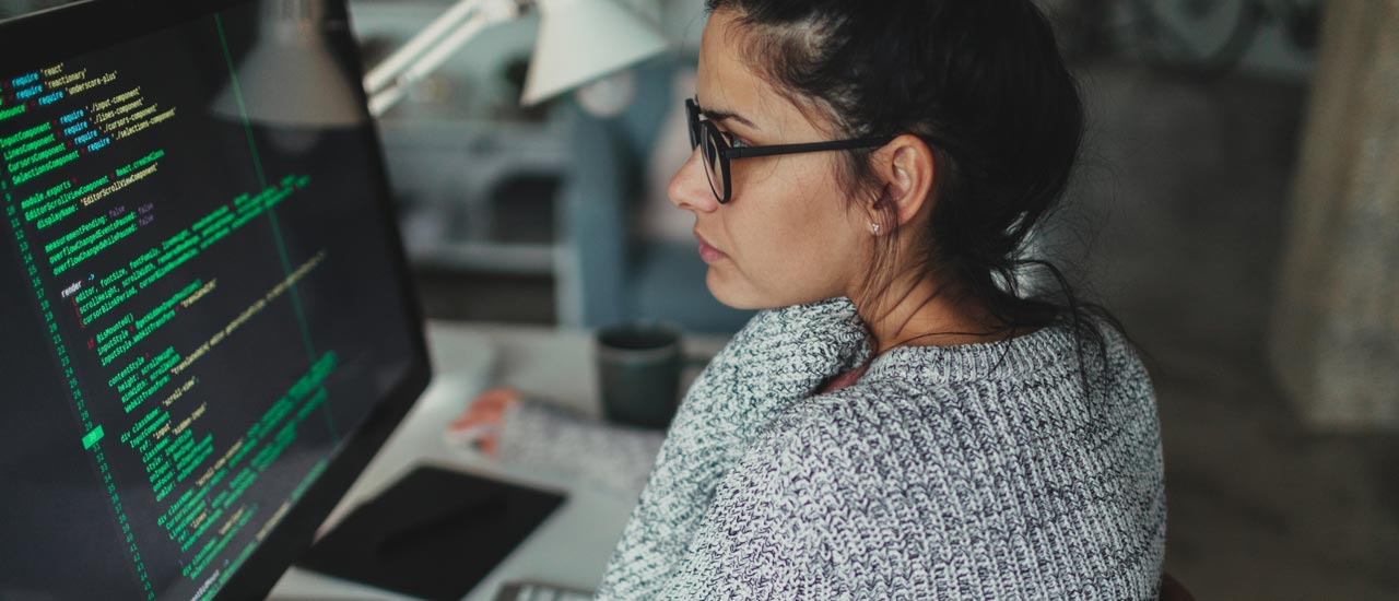 Woman in glasses and gray sweater reviewing code on her computer screen