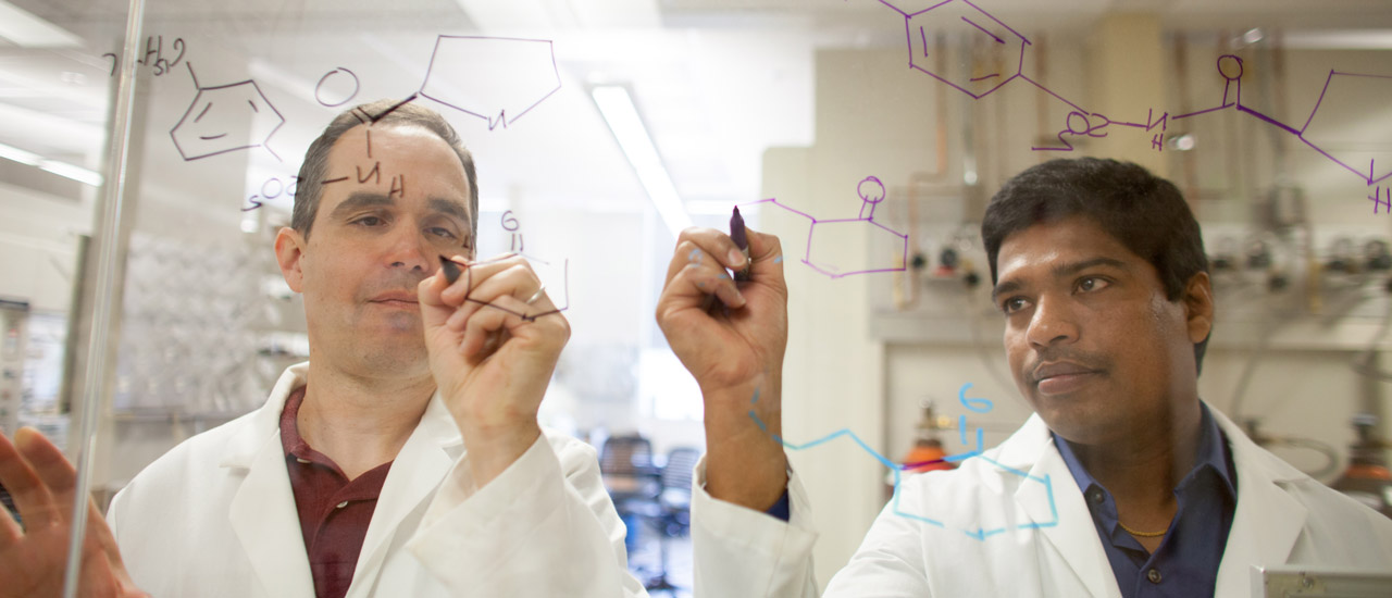 Two chemists in lab coats write chemical sequences with dry erase markers on a clear window