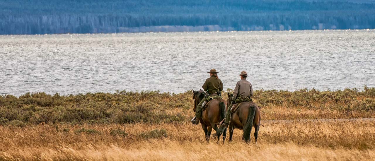 Two park rangers on horseback ride through tall grass, heading towards a large body of water with distant forested hills in the background