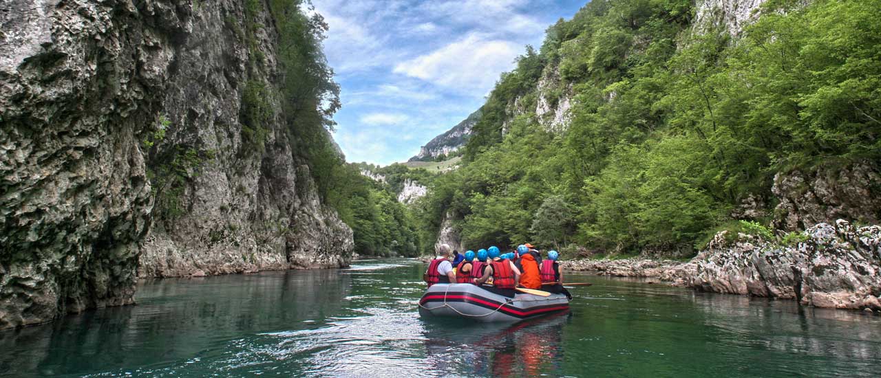 A group of people sitting in an inflatable river raft floating on calm green water through a canyon