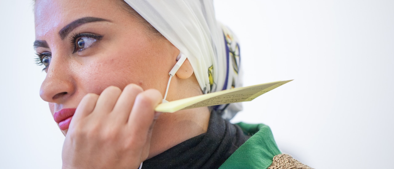 A close up of a woman wearing a head scarf and ear buds looks into the distance while holding notecards in her left hand.