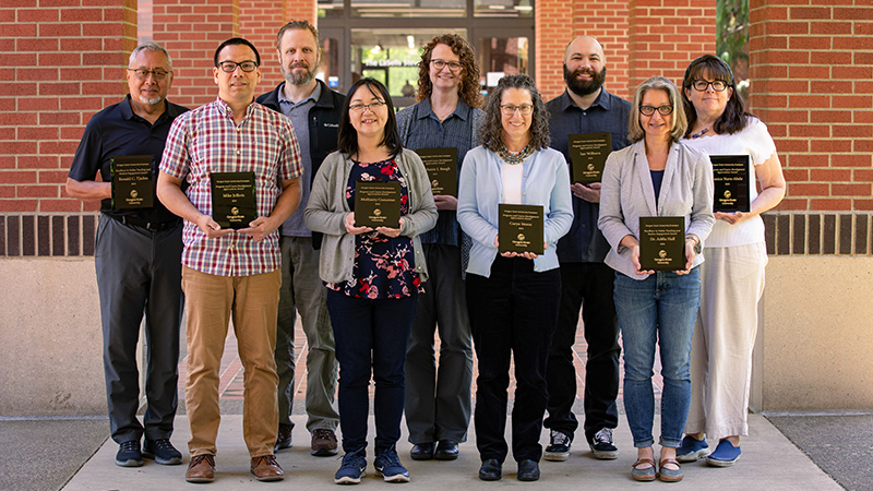 Nine Oregon State University faculty members and support employees stand in an outdoor breezeway while holding award plaques.