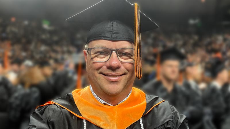 A man wearing a black graduation cap and gown smiles in a selfie in an arena filled with other graduates.