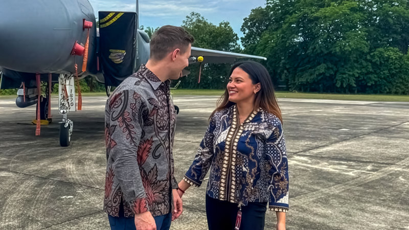 A man and woman, Alya Birzana Devi, in patterned clothing stand smiling on an airstrip in front of a military aircraft.
