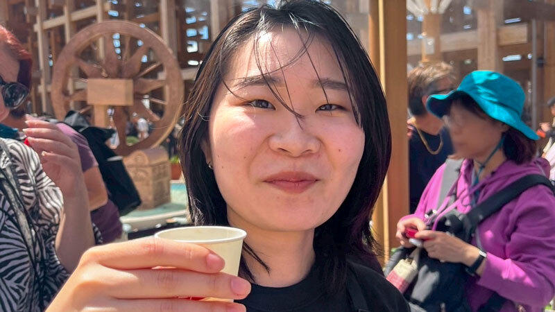 Oregon State University student Asumi Suzuki smiles while holding a paper cup outdoors, with other people and a decorative wooden structure in the background.