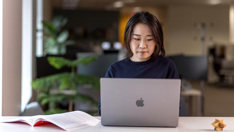 A woman is seated at a desk using a silver laptop in a modern office environment. An open book lies to the left on the desk.