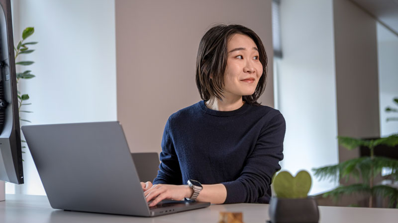 A woman sits at a desk using a laptop, smiling and looking to her left. A small heart-shaped plant is on the desk in front of her.