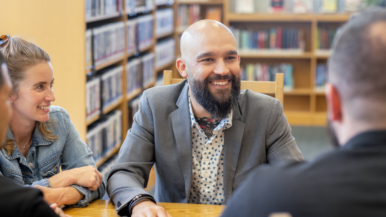 A man sits smiling at a table inside a library while chatting with others.