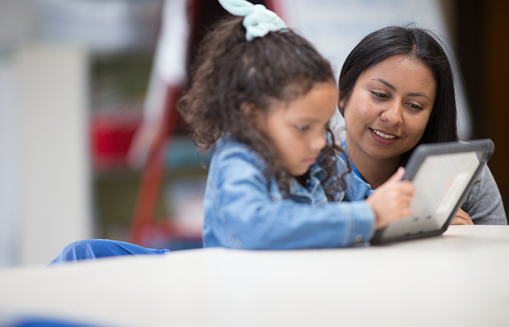 Brenda Contreras is kneeling next to a student in the elementary school classroom where she teaches as part of Oregon State's Master of Arts in Teaching program. The child wears a blue denim jacket and has long, dark and curly hair. Both are looking at a learning tablet that the student is holding.