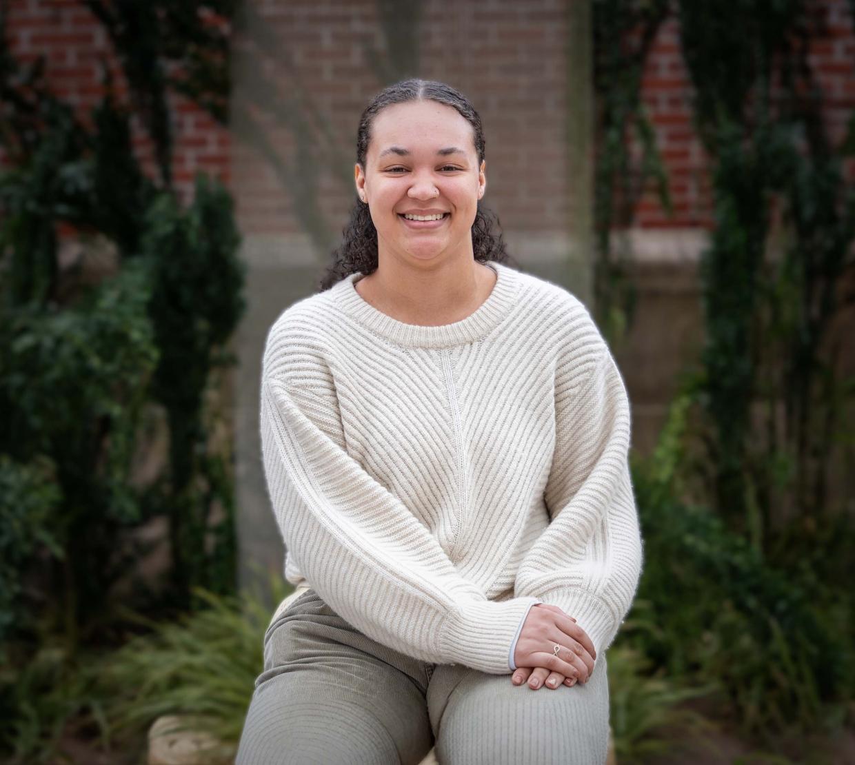 Camrie Smith sits with her hands in her lap wearing a white sweater. She is outdoors with a wall of vines behind her. Her natural hair is pulled back and she smiles broadly at the camera.