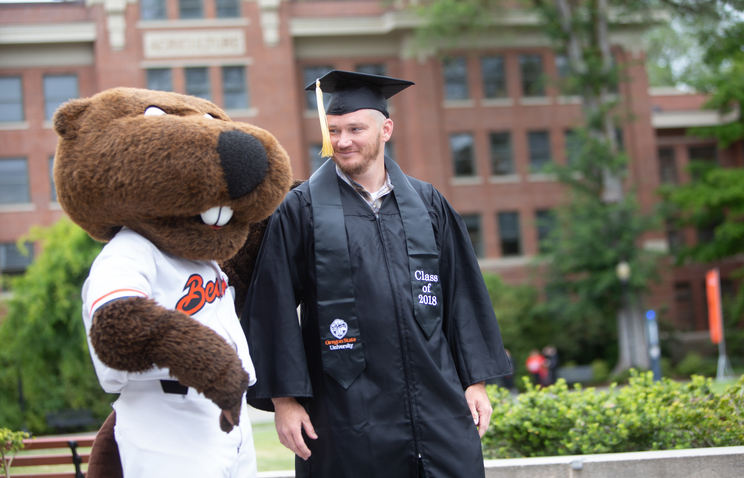 Chris Holt wears his black graduation cap and gown and a black "Class of 2018" stole. Oregon State's mascot Benny the Beaver stands on Chris' left side, wearing a white Beavers jersey.