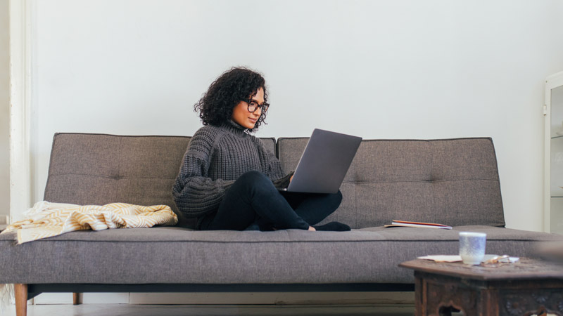 A woman sits cross-legged on a couch while working on a laptop.