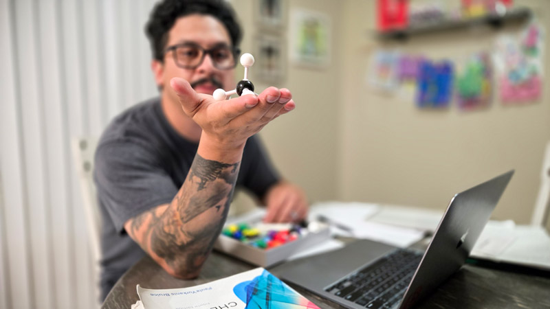 A man sits at a table holding up a molecular model in his hand, with a laptop, textbook and chemistry modeling kit in front of him.