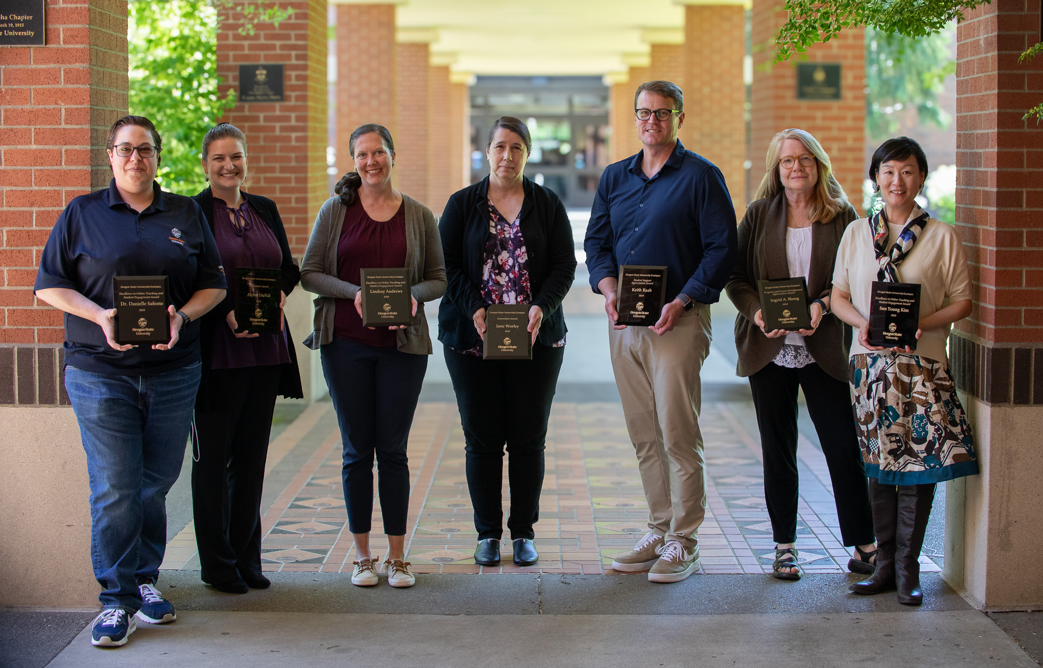 A group of seven individuals stands in a row outdoors under a covered walkway, each holding a small black plaque with their faculty forum awards