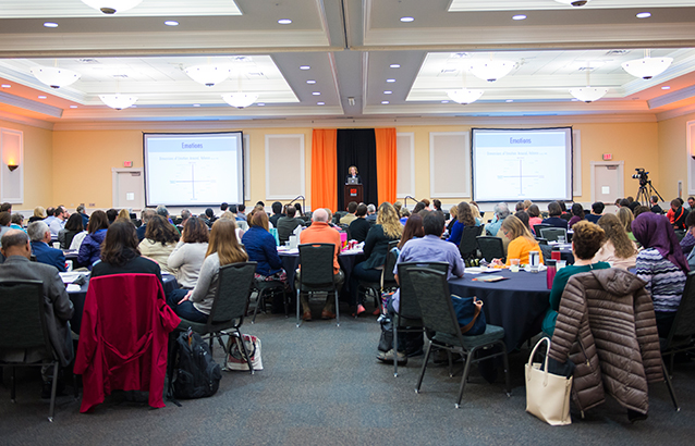 A room full of people seated at round tables listen to keynote speaker Jan Plans at the 2016 Faculty Forum.