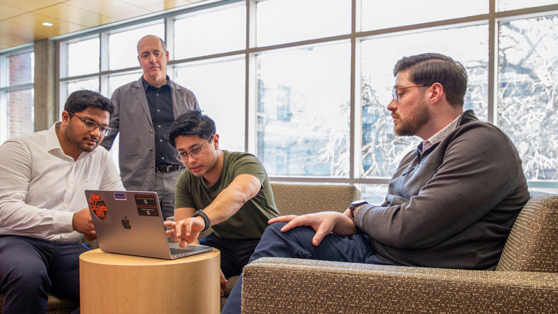 Three Oregon State University engineering students sit in chairs with a professor standing behind them as they look at data on a laptop in a window-filled room.