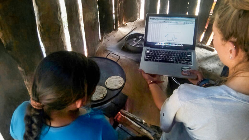 Two people sit in a small hut, looking at a laptop while a round bread cooks on a rustic pan.