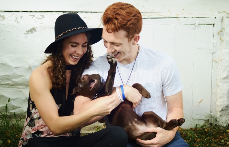 Hannah Thelen is seated outdoors with her husband to her right. The two are holding a puppy with brown fur, which lays on its back in their arms and looks playful with its mouth open and one front paw in the air.