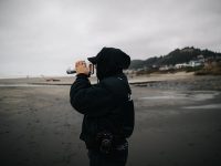 Hooded figure on a beach holding a camera