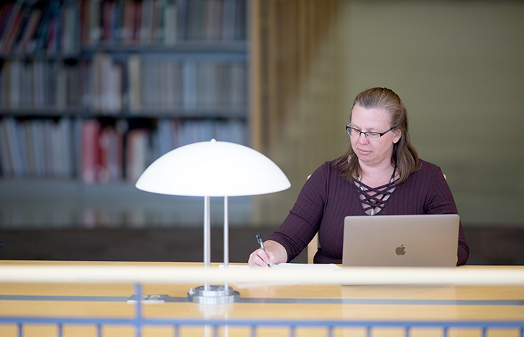 Jennifer Oliphant, a student in Oregon State University's women, gender and sexuality studies program, sits at a desk with a silver laptop open in front of her. She is writing in a notebook to the right of the laptop. In the room, there are several tall bookshelves and a bright white lamp.