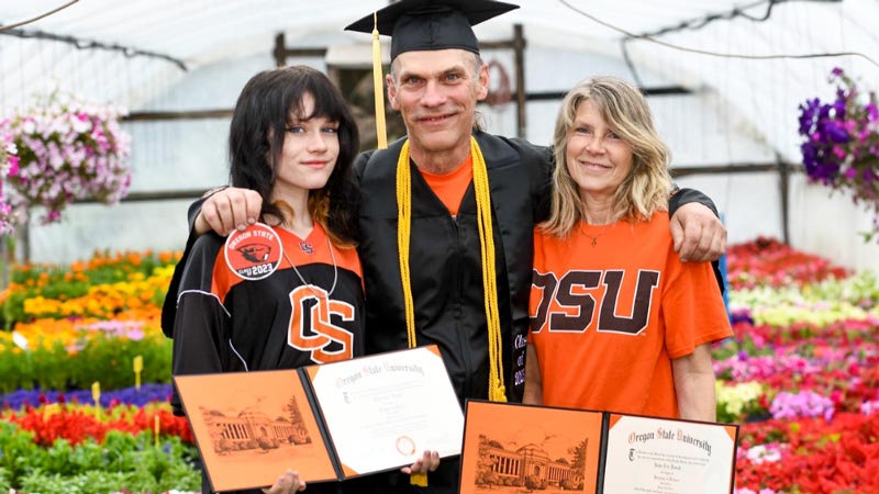 John Dusek in a graduation cap and gown with his wife and daughter presenting his dual degrees to him.