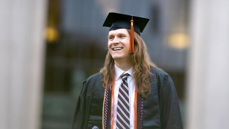 Ecampus grad, Josh Misko smiles toward someone off-camera while wearing his graduation regalia.