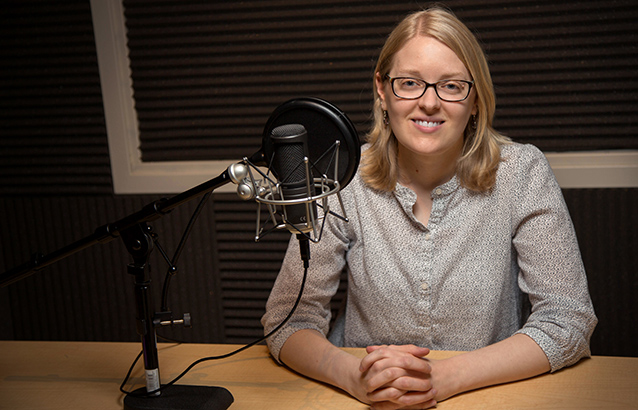 Katie Linder smiles with her hands resting on the table in front of her with her fingers interlocked. On the table is a black microphone with a pop filter.