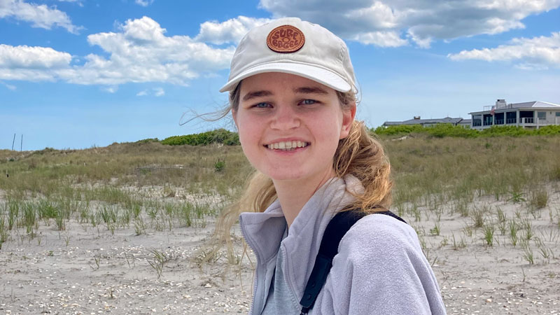 Oregon State University student Lily Vanwingerden stands outdoors in a sandy coastal environment, with scattered grasses and shells nearby.