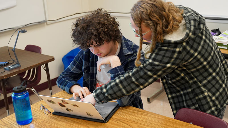 Two individuals in a secondary education classroom setting, one seated and focused on a laptop, while the other stands, pointing at the screen.