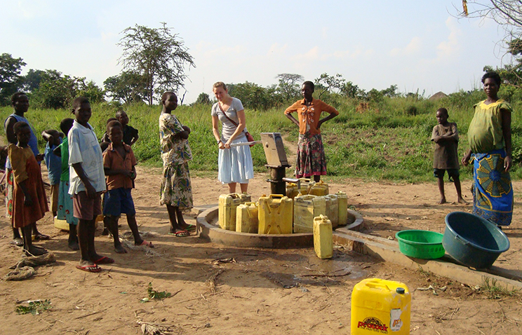 A group of people stand around a water pump that is surrounded by large yellow jugs. One person is pumping water into one of the jugs.