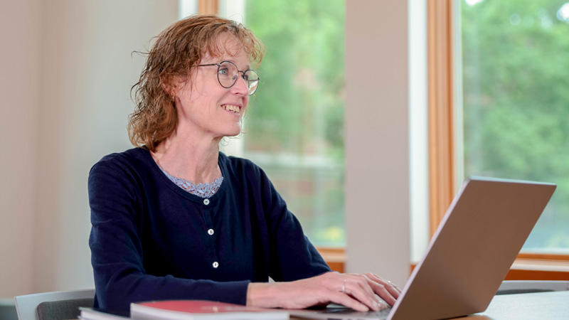 A woman wearing glasses sits while working on a laptop, with a book on the table in the foreground.