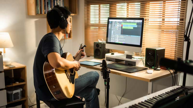 A person wearing headphones sits on a stool at a desk while playing guitar and looking at a computer screen.