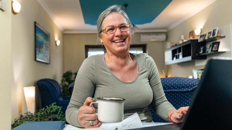 A middle aged woman wearing glasses smiles at a laptop while holding a mug.