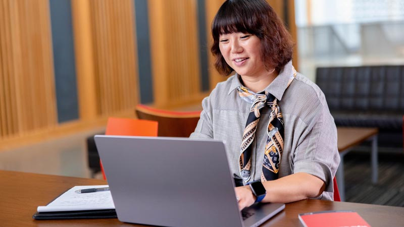 Ecampus student Karen Baek sits at a laptop in an OSU building. She has a book and handwritten notes nearby.