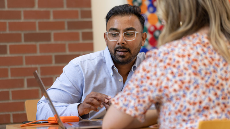 An Oregon State computer science student points at a computer screen while talking to someone sitting across the table