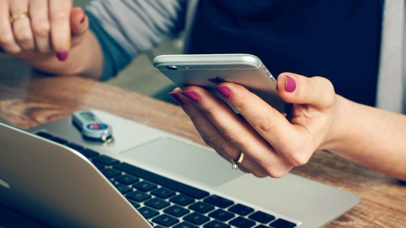 A closeup of someone sitting at an open laptop with a phone in their hand