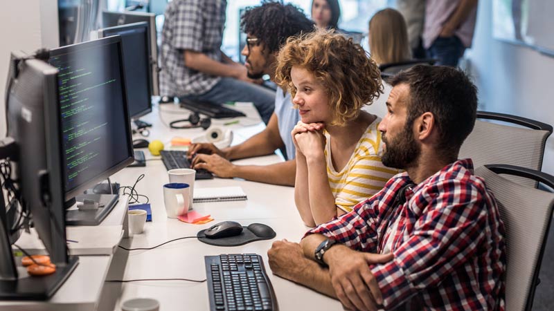 A photo of two people working at a desk and looking at a computer.