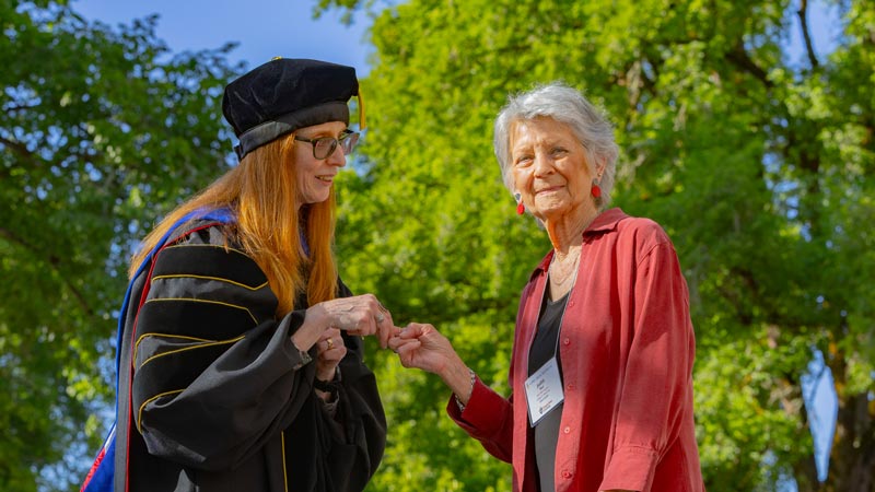 Oregon State University graduate Judith Paul shakes hands with Benita Blessing outdoors, with large green trees behind them.