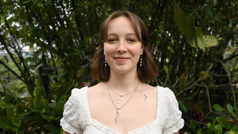 A smiling woman stands outside in front of a large green bush.