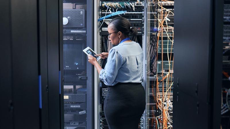 A woman stands working inside a server room, positioned between tall black server racks. She is holding and examining a digital tablet.