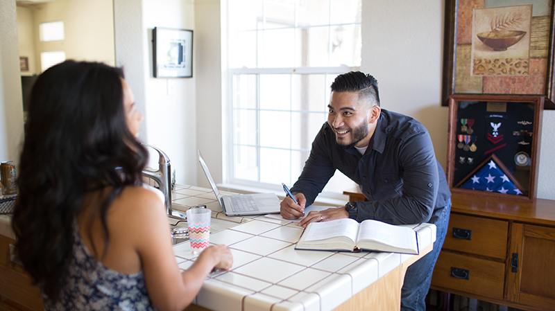 A smiling man leans on a kitchen counter with a textbook and notepad while talking to his wife in the foreground.