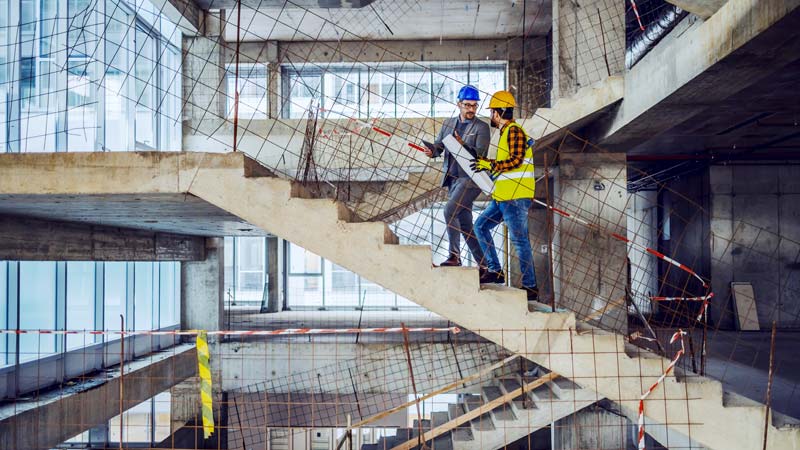 Two people wearing hardhats and construction vests walk up stairs in a building under construction.
