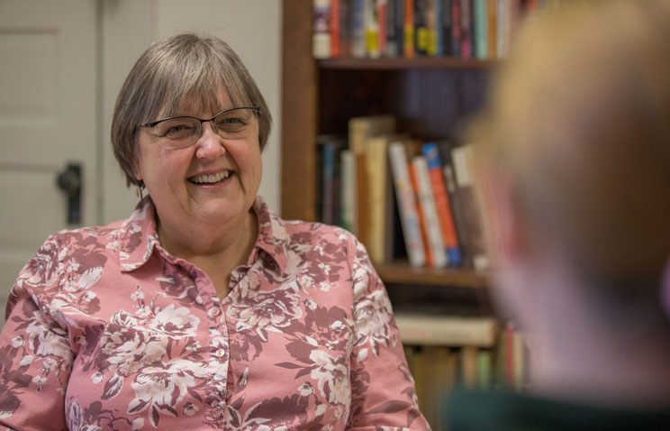 Brenda Kellar, Ecampus anthropology advisor. Brenda wears a pink floral patterned button-up shirt and has short gray hair. She is seated in front of a bookshelf across from a person to whom she is speaking.