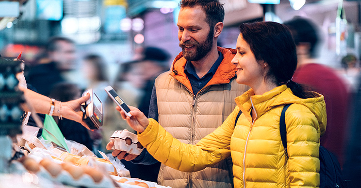 Two shoppers purchase eggs at an outdoor market, with a woman holding a smartphone as she scans a hand-held payment device