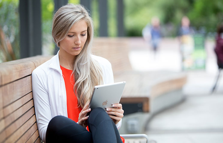 Ecampus alumna Calais Bickford-St. Pierre sits sideways on a long, curved wooden bench. She is looking down at a silver tablet that rests against her knees.