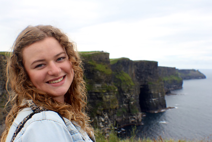 Carlee Sedgwick, a sustainability and sociology double major with Oregon State University Ecampus, stands with ocean cliffs behind her. She is in the environmental and natural resource option in the sociology bachelor's degree program.